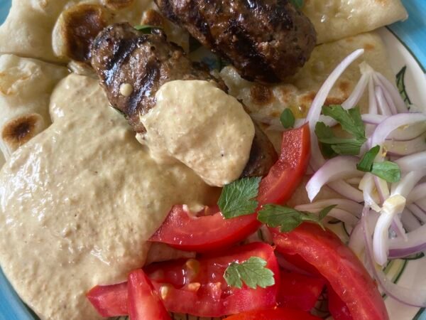 Plato de kebab estilo Thanasis servido con pan pita artesanal, salsa cremosa de pimentón rojo, rodajas de tomate fresco y cebolla morada, en un plato azul claro.