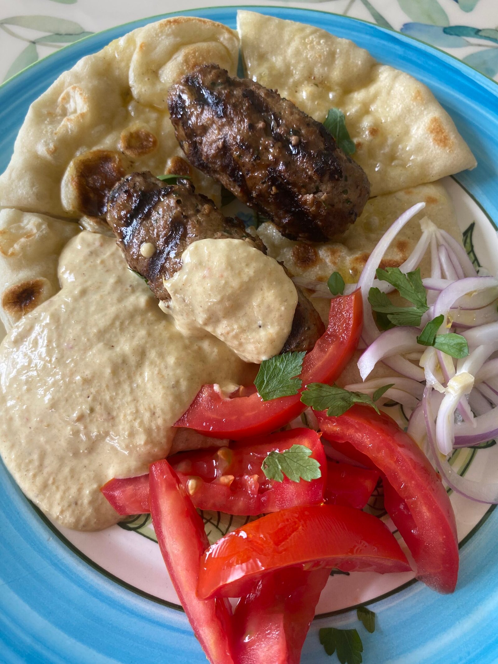 Plato de kebab estilo Thanasis servido con pan pita artesanal, salsa cremosa de pimentón rojo, rodajas de tomate fresco y cebolla morada, en un plato azul claro.